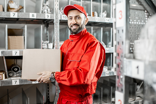Handsome warehouse worker in red uniform taking some products from the shelves at the storage with car parts