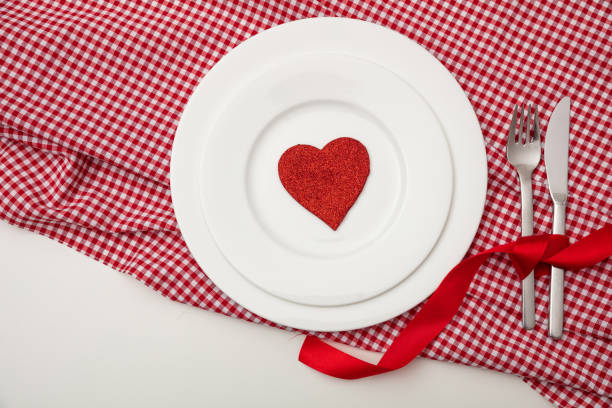 Valentines day dinner, table, place setting. Red heart on white plates, red checkered table cloth, white background, copy space, top view.