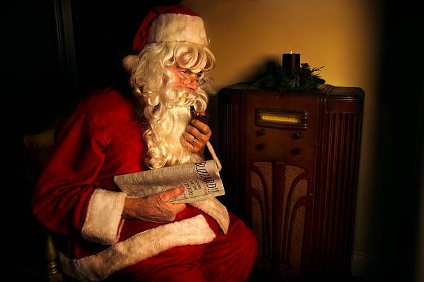 santa claus sentado por radio de antigüedades y sosteniendo un periódico - retro christmas audio fotografías e imágenes de stock