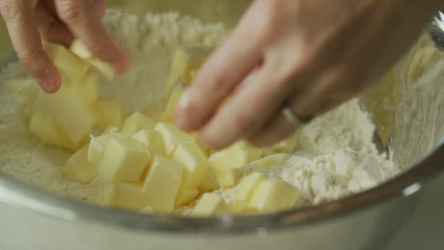 A Young Woman's Hands Cut Butter into Small Pieces with a Large Kitchen Knife on a Wooden Cutting Board and then Pick Up the Butter and Sprinkle It Over Flour in a Metal Mixing Bowl