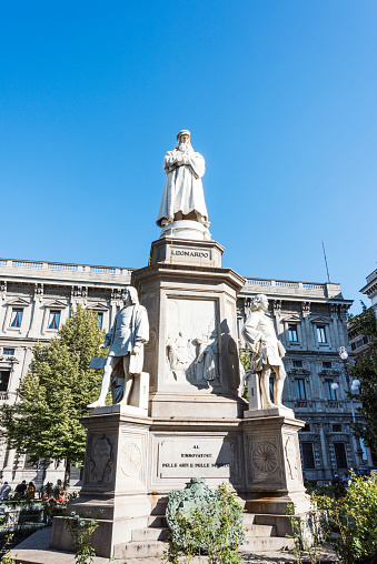 Milan, Italy - August 27, 2018: Monument of Leonardo da Vinci, Piazza della Scala, Milan Italy. The monument is in the center of square and was designed by sculptor Pietro Magni (1872). Teatro alla Scala opera house occupies the north-western side of the square. Green bushes, flowers, historic buildings and blue sky with clouds are in the image.