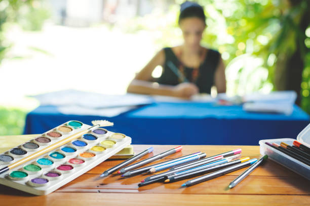 Woman blurred in the background, painting and drawing, upon a table, and some pencils and ink focused in the foreground.