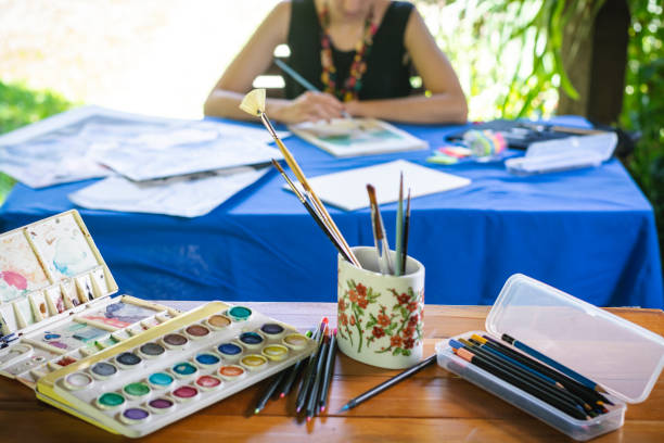 Woman blurred in the background, painting and drawing, upon a table, and some pencils and ink focused in the foreground.