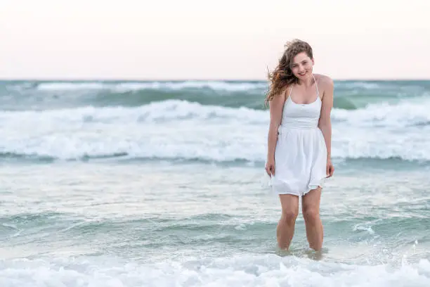 Young happy woman in white dress on beach pink sunset in Florida panhandle with wind, ocean waves, standing similing in water