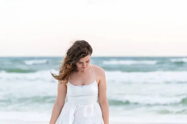 Young sad woman in white dress on beach sunset, dusk, twilight in Florida panhandle with wind, ocean waves, tan skin, looking down