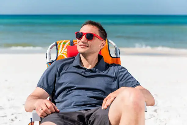 Young man millennial in red sunglasses on beach chair during sunny day in Florida, lying reclining back, ocean horizon, white sand, gulf of mexico