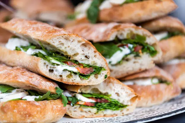 Closeup of fresh display of stacked pile of panini bread, mozzarella melted cheese, vegetarian italian tomatoes, basil lettuce in store, shop, cafe buffet catering sandwiches