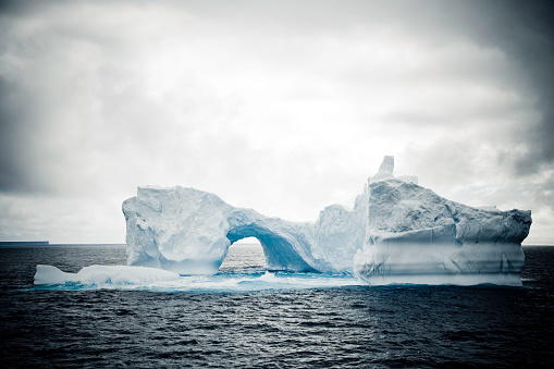 amazing shaped iceberg in the antarctica with natural arch under cloudy sky.
