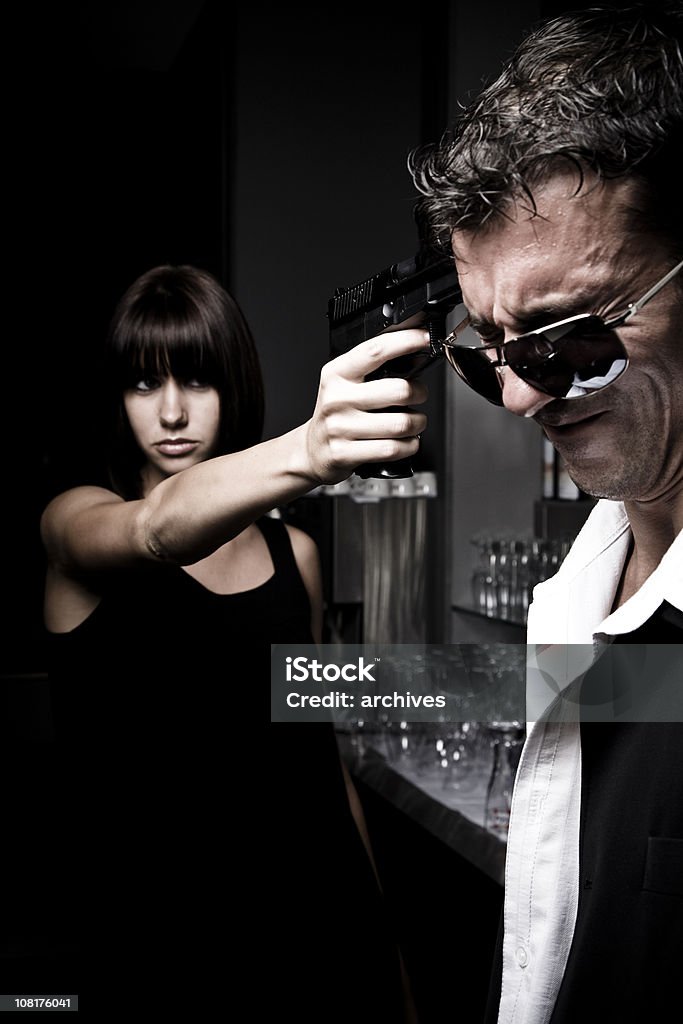 Young Woman Pointing Gun at Head of Grimacing Man  Dark Stock Photo