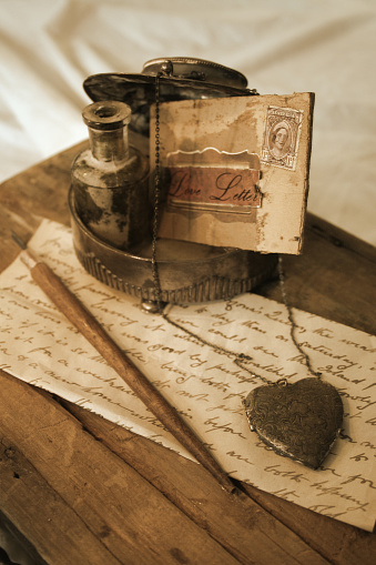 A narrative composition of antique objects: ink bottle, quill, hand-written letter, silver locket and stamped envelope.  Desaturated and sepia toned