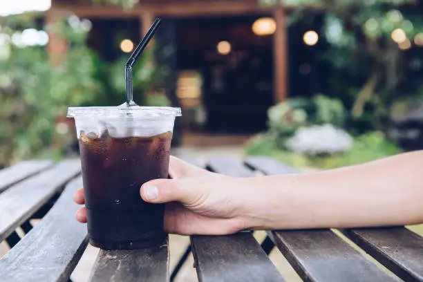 Photo of Cropped shot of someone hand touching a plastic cup of iced Americano (black coffee) on the wood plank table.