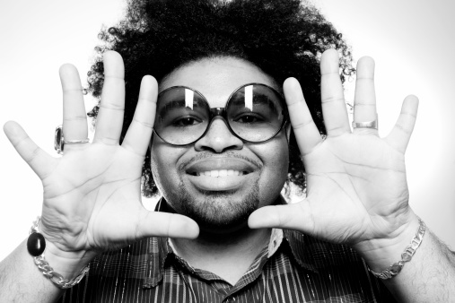 Portrait of a man with big hair and big glasses framing his face with his hands.