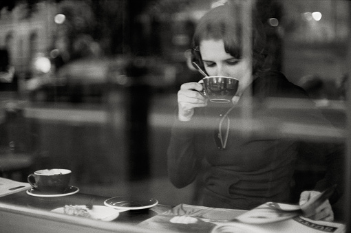 Woman drinking coffee and reading magazine behind the window of a coffee house.