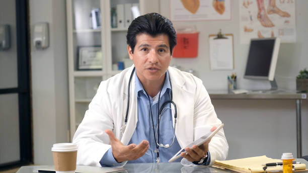 Male doctor shares advice during web conference A mid adult male doctor sits behind a desk in his office and gestures as he shares advice during a web conference. He is holding a digital tablet. doctor utensils stock pictures, royalty-free photos & images