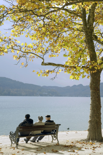 Three senior men sitting in sunlight on park bench under plane trees at water´s edge of mountain lake in autumn. Location: Lake Wörthersee, City of Klagenfurt, Austria.