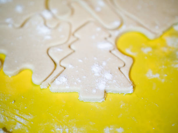 eine nahaufnahme von einem weihnachtsbaum-ausschnitt und andere formen der urlaub in rohzucker cookie-teig auf ein gelbes schneidebrett mit mehl bestreut an der spitze ein festliches hintergrundbild für den urlaub machen. - candy cane christmas cookie raw food stock-fotos und bilder