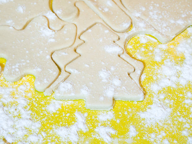eine nahaufnahme von einem weihnachtsbaum-ausschnitt und andere formen der urlaub in rohzucker cookie-teig auf ein gelbes schneidebrett mit mehl bestreut an der spitze ein festliches hintergrundbild für den urlaub machen. - candy cane christmas cookie raw food stock-fotos und bilder