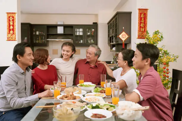 Happy big Asian family sitting at dinner table and celebrating Lunar New Year, couplets with best wishes for coming year in the background