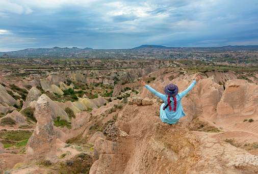 A young girl in turquoise clothes sits on top of a rock in Cappadocia and looks spread her arms out on mountains, ravines and a blue sky.