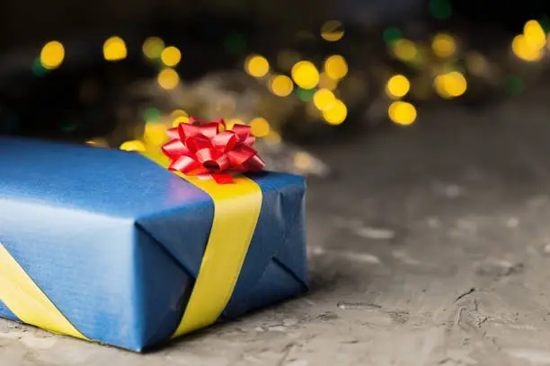 Gift in blue box with yellow ribbon and red bow, against yellow bokeh background.