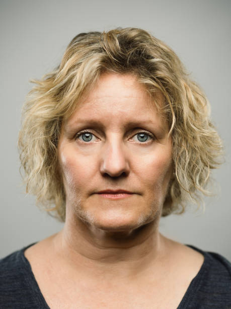 Real caucasian adult woman with blank expression Close up portrait of caucasian mature woman with blank expression against gray background. Vertical shot of real british woman staring in studio with curly blond hair and green eyes. Photography from a DSLR camera. Sharp focus on eyes. fine art portrait photos stock pictures, royalty-free photos & images