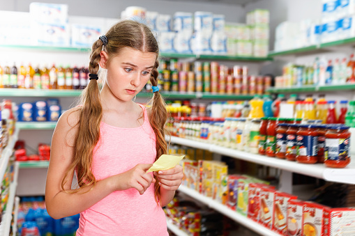 Positive  glad  cheerful smiling tween girl choosing food products on shopping list in supermarket