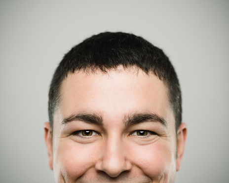 Close-up portrait of real young man looking at camera and smiling. Happy caucasian male with brown hair against white background. Horizontal studio photography from a DSLR camera. Sharp focus on eyes.