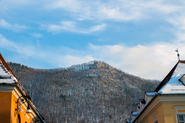 A cluster of rural houses sits at the base of a frosted hillside, illuminated by warm winter sunlight. Snow-covered roofs and bare trees create a peaceful seasonal atmosphere, contrasting with the golden tones of the buildings. The scene captures the quiet charm of a small village in winter.