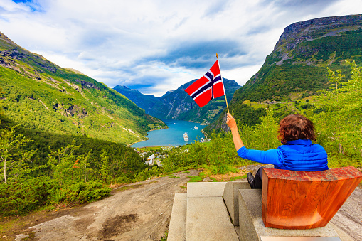Tourism vacation and travel. Female tourist enjoying beautiful view over magical Geirangerfjorden from Flydalsjuvet viewpoint, holding norwegian flag.