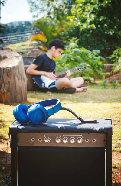 Young adult, sitting on the floor and playing guitar, unfocused in the background and a headphone and a speaker focused in the foreground.