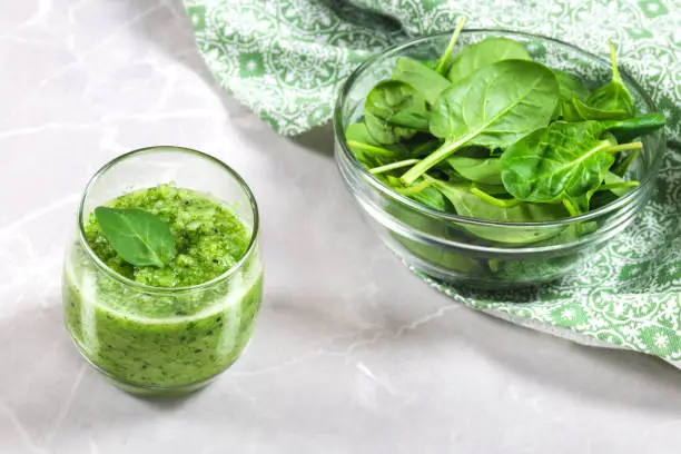 Green healthy smoothie in a cup decorated with a spinach leaf and a bowl of fresh spinach in the background.