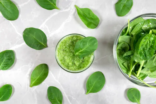 Green healthy spinach smoothie in a  glass, bowl with fresh spinach and spinach leaves on a marble background.