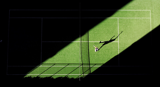 A man plays an overhead smalsh shot in a shaft of light across a tennis court\n