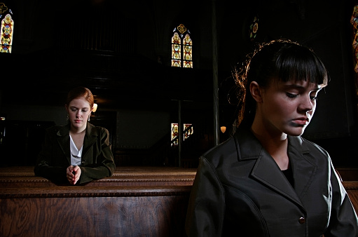 Two women in business outfits sitting and kneeling in a dark, old, cathedral.  Horizontal with copy space.