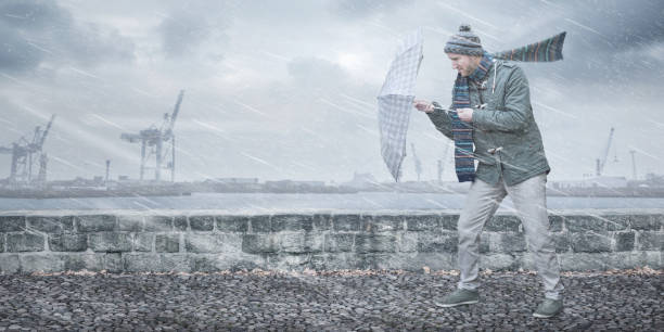 Man with an umbrella is facing strong wind and rain A pedestrian is walking down a footpath on a dockside. He is opposing a strong wind and rain with an umbrella. In the background you can see a port area with cranes. gale stock pictures, royalty-free photos & images