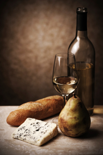 Moody lighting on still life of white wine, bleu cheese, pear, bread, with subtle, subdued tones.