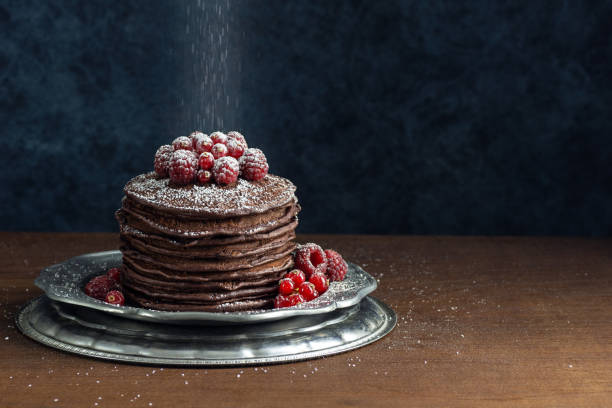 Powdered Sugar Sprinkling on Dark Chocolate Pancakes with Red Currants and Raspberries Powdered sugar is being sprinkled from above a tall stack of dark chocolate pancakes. The pancakes are on a silver metal plate with a garnish of raspberries and red currants. Copy space on the dark blue background. sprinkling powdered sugar stock pictures, royalty-free photos & images