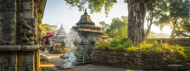 Smoke swirling through the sunlit foliage shading the ancient shrines of Gorakhnath Temple at the UNESCO World Heritage Site of Pashupatinath in the heart of Kathmandu, Nepal's vibrant capital city.