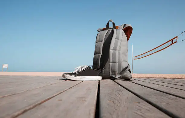 Summer vacation concept. Backpack and sneakers are on the wooden desk near the beach and sea.Blue clear sky and sun