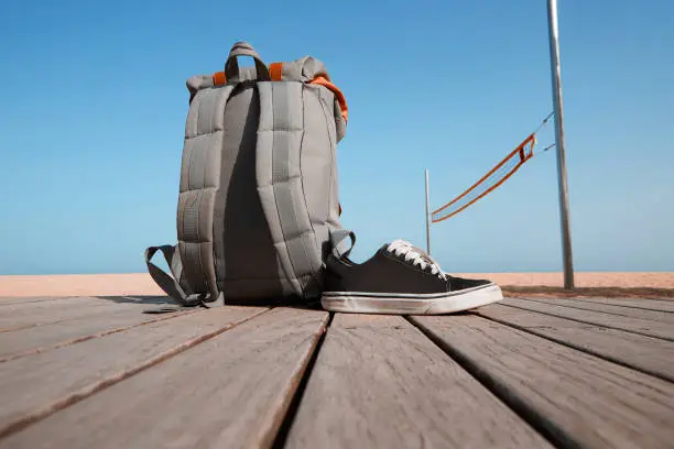 Summer travel concept. Backpack and sneakers are on the wooden desk near the beach and sea.Blue clear sky and sun