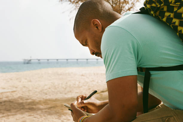 istock Pensive African-American hipster man wearing backpack and making notes in copybook outside of the city. Student preparing for lesson at college on the beach. 1081982232