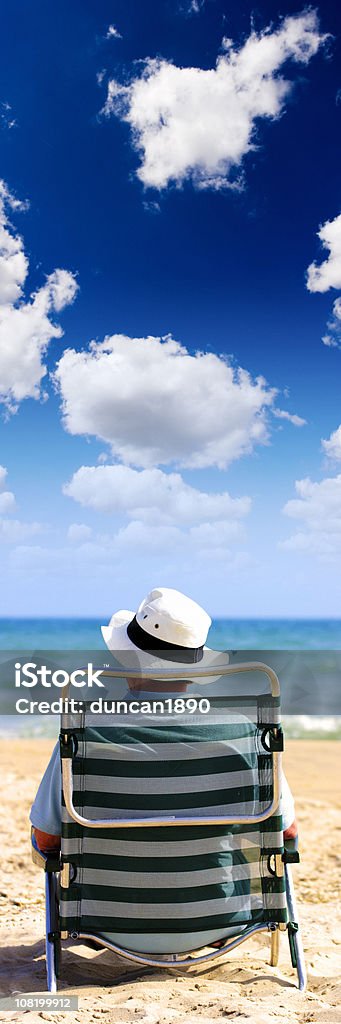 Uomo con cappello seduto su una sedia sulla spiaggia - Foto stock royalty-free di Sedia a sdraio