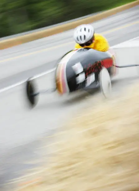 Soapbox derby competitor speeds downhill in crouched racing motion blur.  The Soap Box Derby is a youth car racing program which has been run in the United States since 1934. World Championship finals are held each July at Derby Downs in Akron, Ohio. Race cars are unpowered, relying entirely upon gravity to move downhill.  Early cars were constructed of wood from crates and other scrap sources, with wheels from baby buggies and wagons.  Today, using standardized wheels with precision ball bearings, modern racers start on a ramp at the top of a hill, and attain speeds of up to 35 miles per hour.  Irondequoit, New York, 2008.