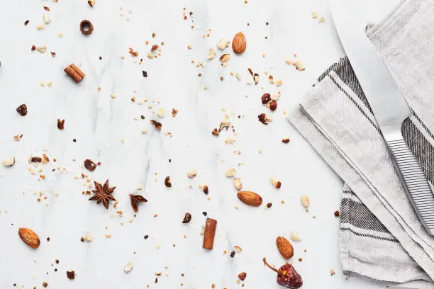 Baking background for baking cookies or cakes. Knife and dishtowel with cinnamon, anise, hazelnut, almond and dried orange crumbs on white marble table. Top view.