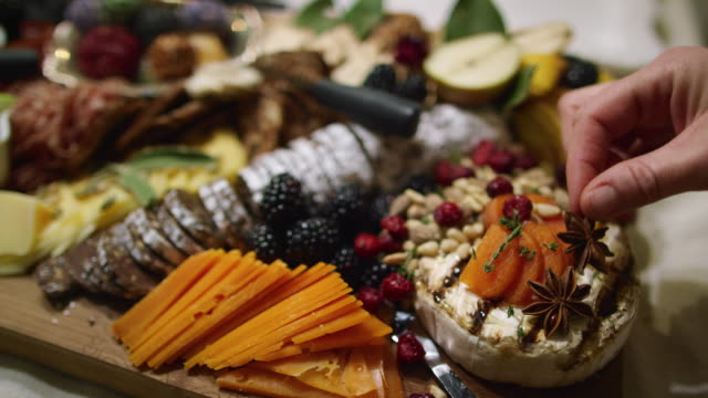 A Woman's Hand Places Star Anise on an Appetizer Charcuterie Meat/Cheeseboard with Various Fruits, Sauces, a Chocolate Log, and Garnishes on a Table at an Indoor Celebration/Party