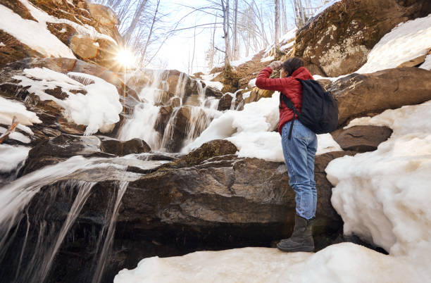 Beautiful girl in red jacket with black backpack stands arm up in front of the winter waterfall and orange sun