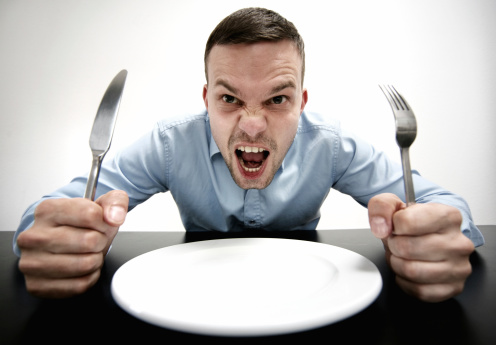 man sitting on a table in front of an empty plate. knife and fork in his hands.he is screaming. he is hungry.