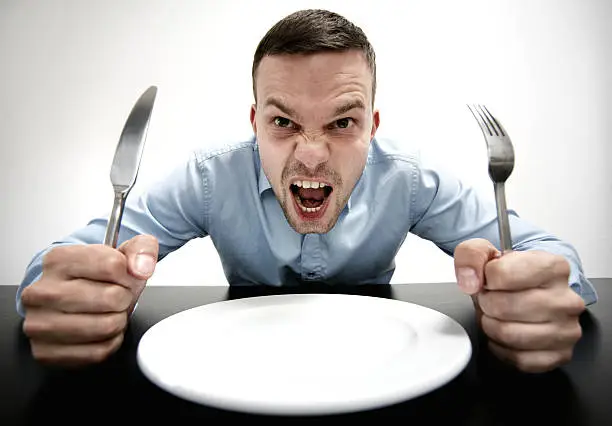 man sitting on a table in front of an empty plate. knife and fork in his hands.he is screaming. he is hungry.