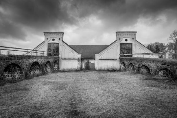 istock Vintage rural scene, dramatic front view of two black and white old rustic worn stone barn building farmhouse with dark sky. Two arched stone bridges in foreground. 1082058068