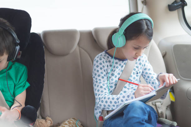 Elementary-age children entertaining themselves on a road trip in the car A young ethnic girl colors and listens to music on a road trip. Her brother is sitting in a booster and watching a movie on a digital tablet. They are both wearing headphones. Distracted stock pictures, royalty-free photos & images